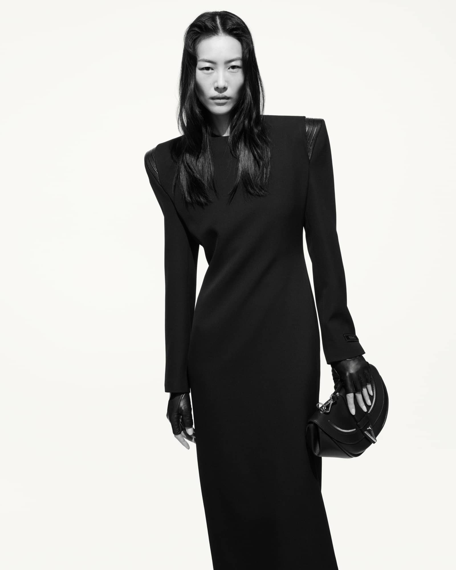 Model in long black dress with structured shoulders holding a black handbag against white background