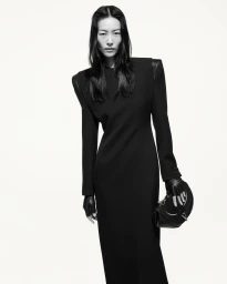 Model in long black dress with structured shoulders holding a black handbag against white background