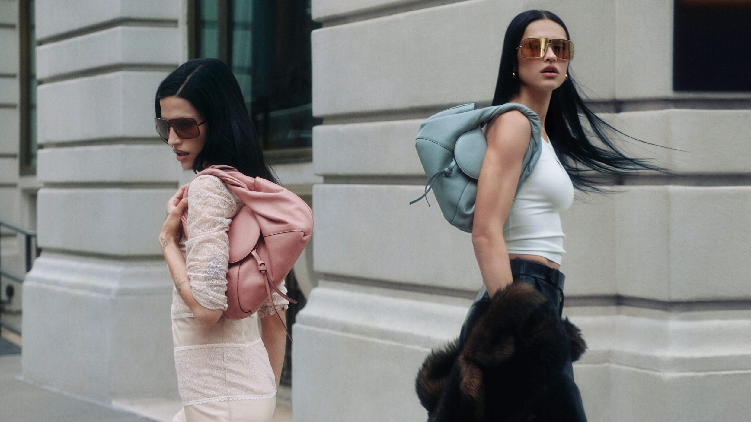Two women with dark hair and sunglasses posing outdoors carrying leather shoulder bags in front of a classical stone building.