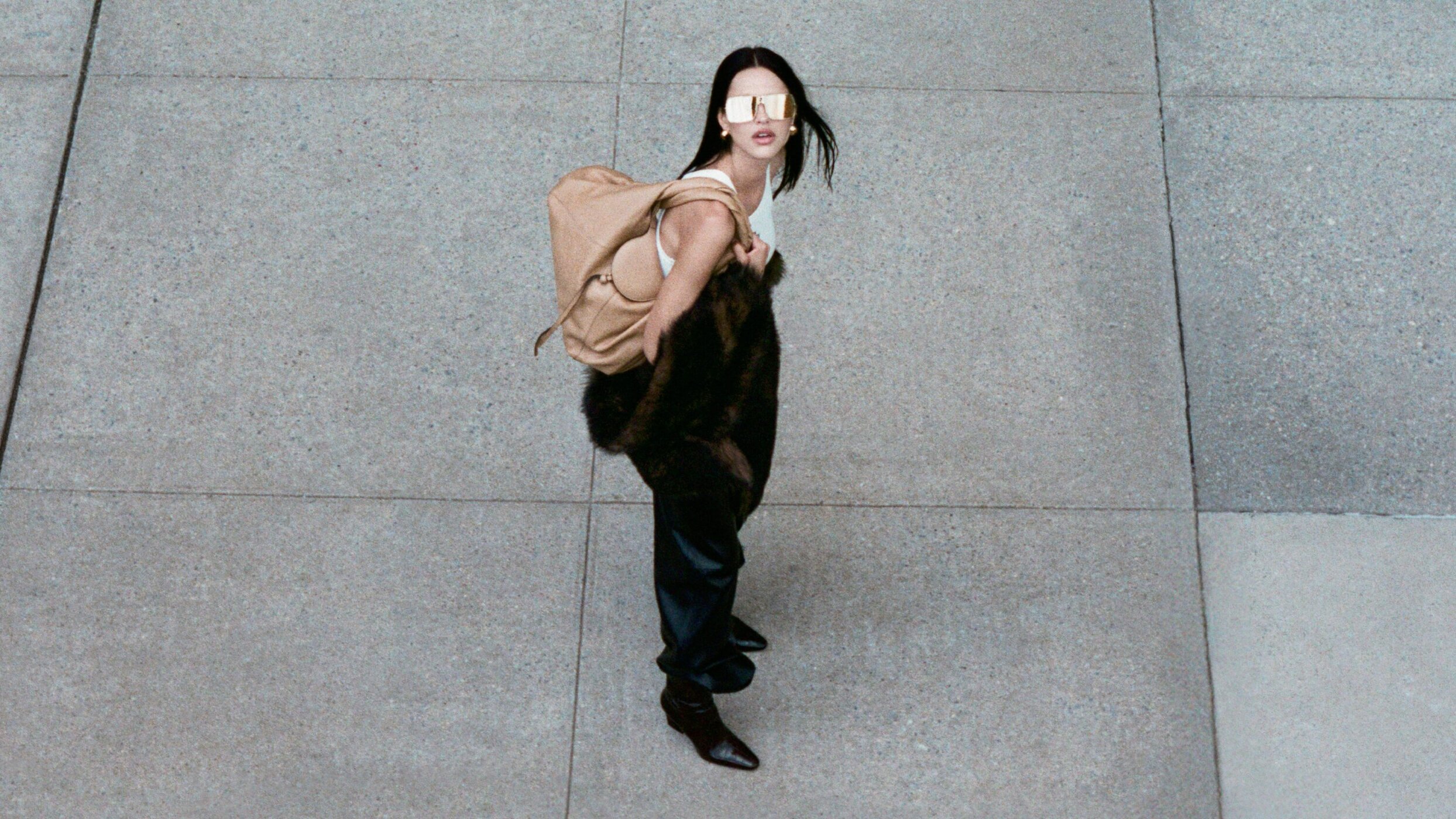Model wearing dark sunglasses and carrying a tan shoulder bag on a concrete pavement.