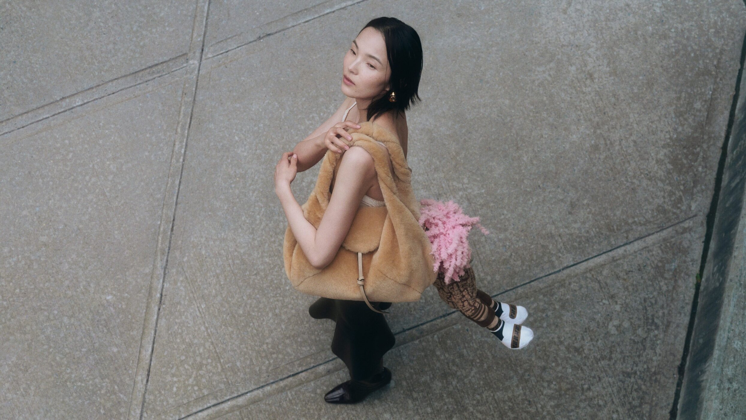Model holding a fuzzy tan shoulder bag while posing against a concrete sidewalk in an overhead angle shot.