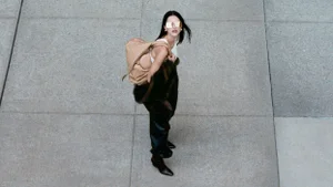 Model wearing dark sunglasses and carrying a tan shoulder bag on a concrete pavement.