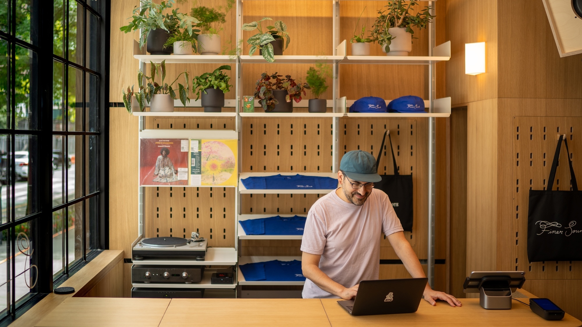 Man wearing a cap and glasses standing behind a wooden counter using a laptop inside a record store with shelving units and potted plants.