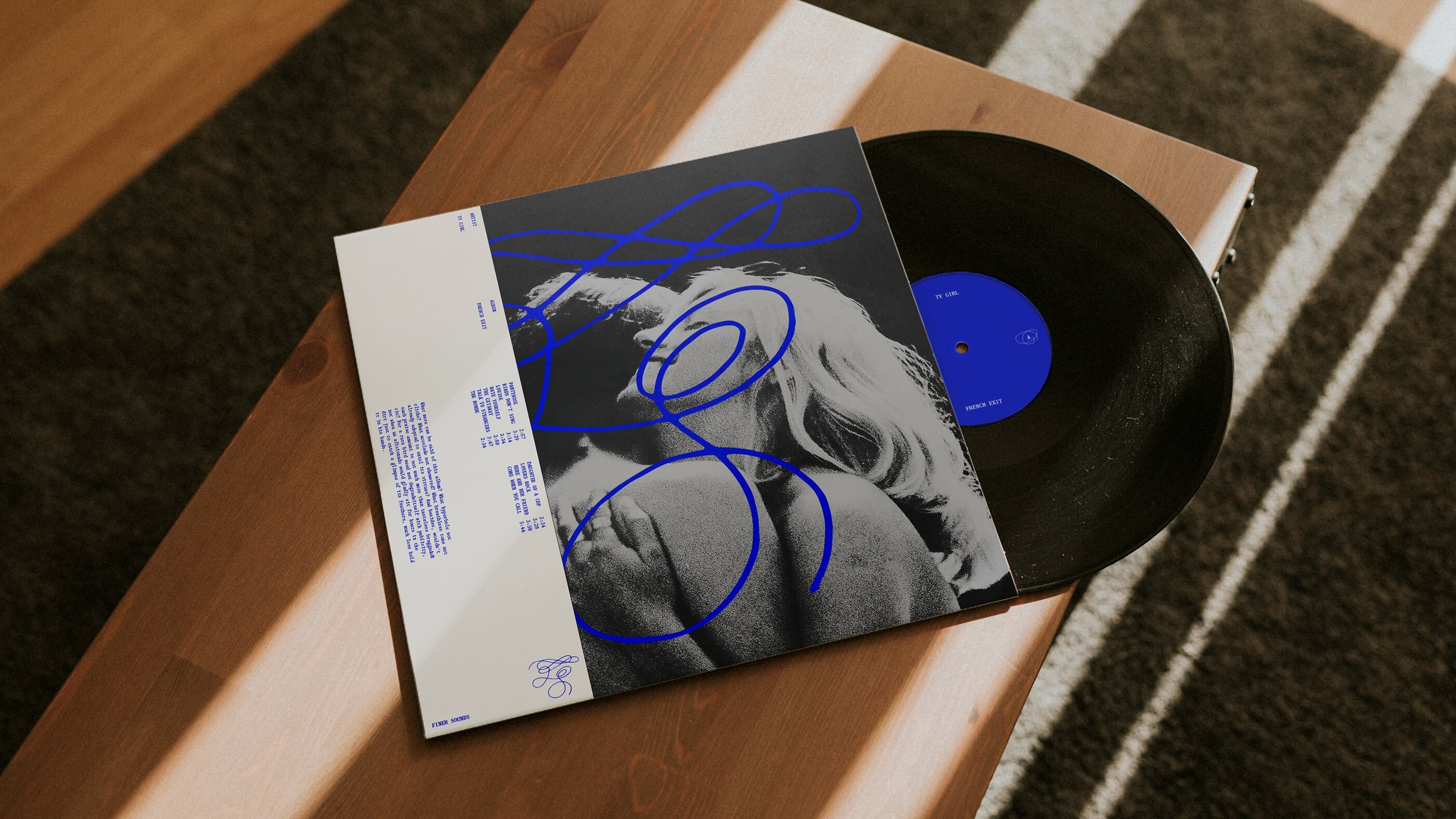 Vinyl record and album cover featuring black and white imagery with blue scribble overlays resting on a wooden surface.