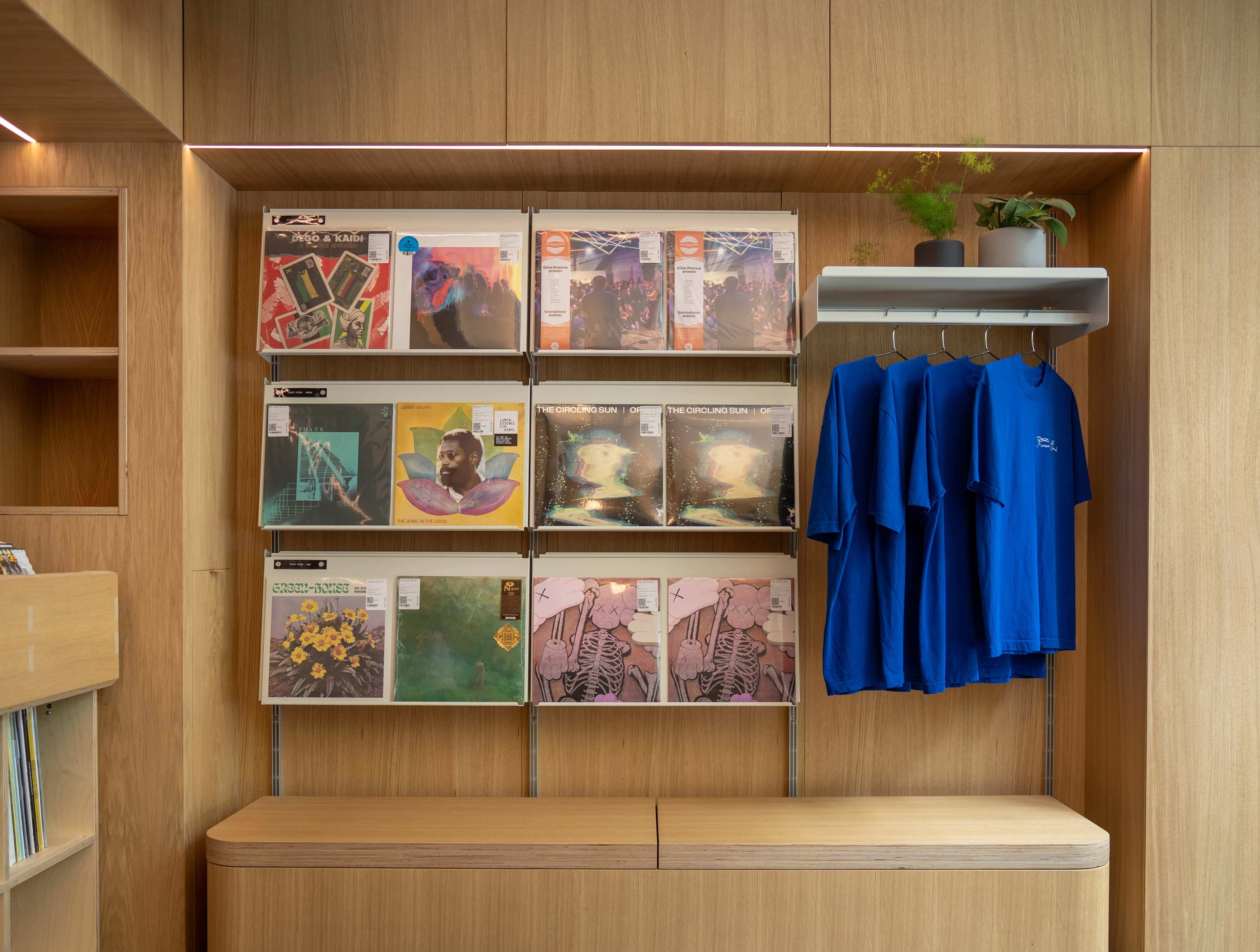 Retail display wall with wooden paneling showcasing vinyl records in shelves and four blue t-shirts hanging on a rack.
