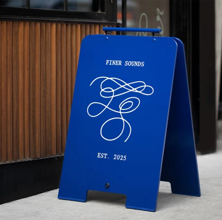 Blue A-frame sign with white branding text and abstract line art standing on pavement outside a building entrance.