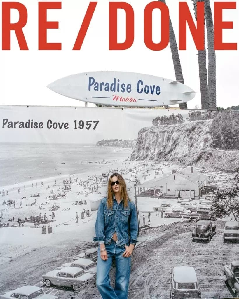 Alessandra Ambrosio wearing a denim jacket and jeans standing before a Paradise Cove backdrop in a RE/DONE campaign photograph.