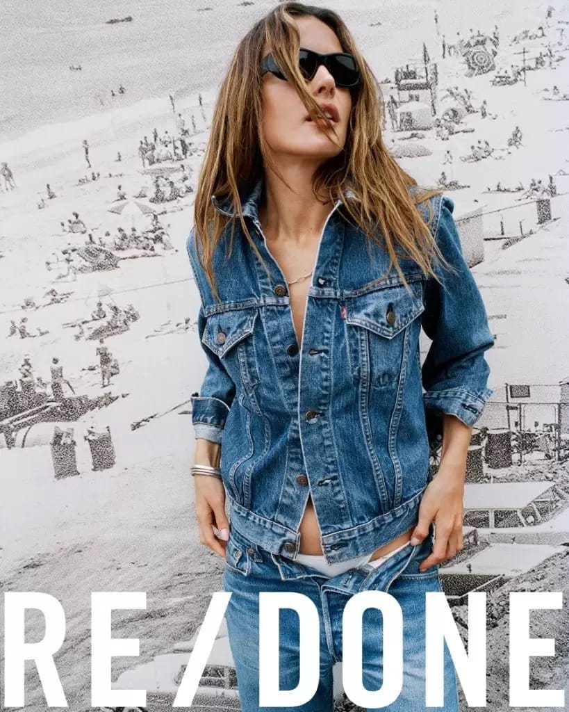 Alessandra Ambrosio in a denim jacket standing against a large black and white beach scene backdrop for RE/DONE.