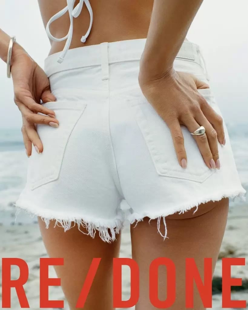 Close-up rear view of a model wearing white denim shorts and a white bikini top outdoors near the ocean.