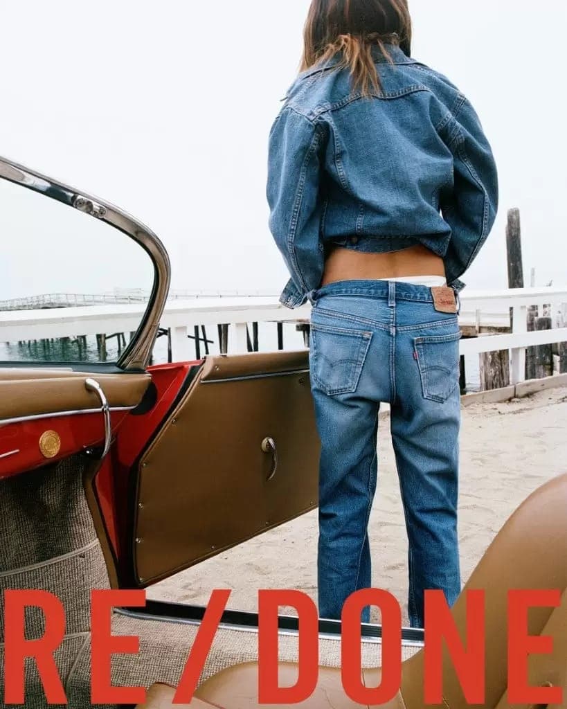 Model standing on a sandy beach wearing a denim jacket and jeans by a red vintage car, photographed by Zoë Ghertner.