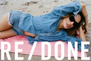 Model Alessandra Ambrosio wearing a denim shirt and shorts lying on a pink beach towel on sand.