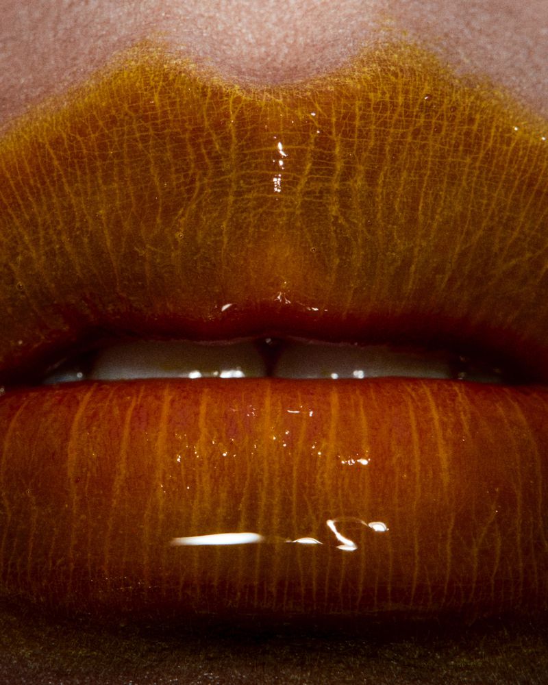 Close-up of an adult female's lips painted with glossy, iridescent amber and gold lipstick, capturing a beauty and fashion aesthetic.