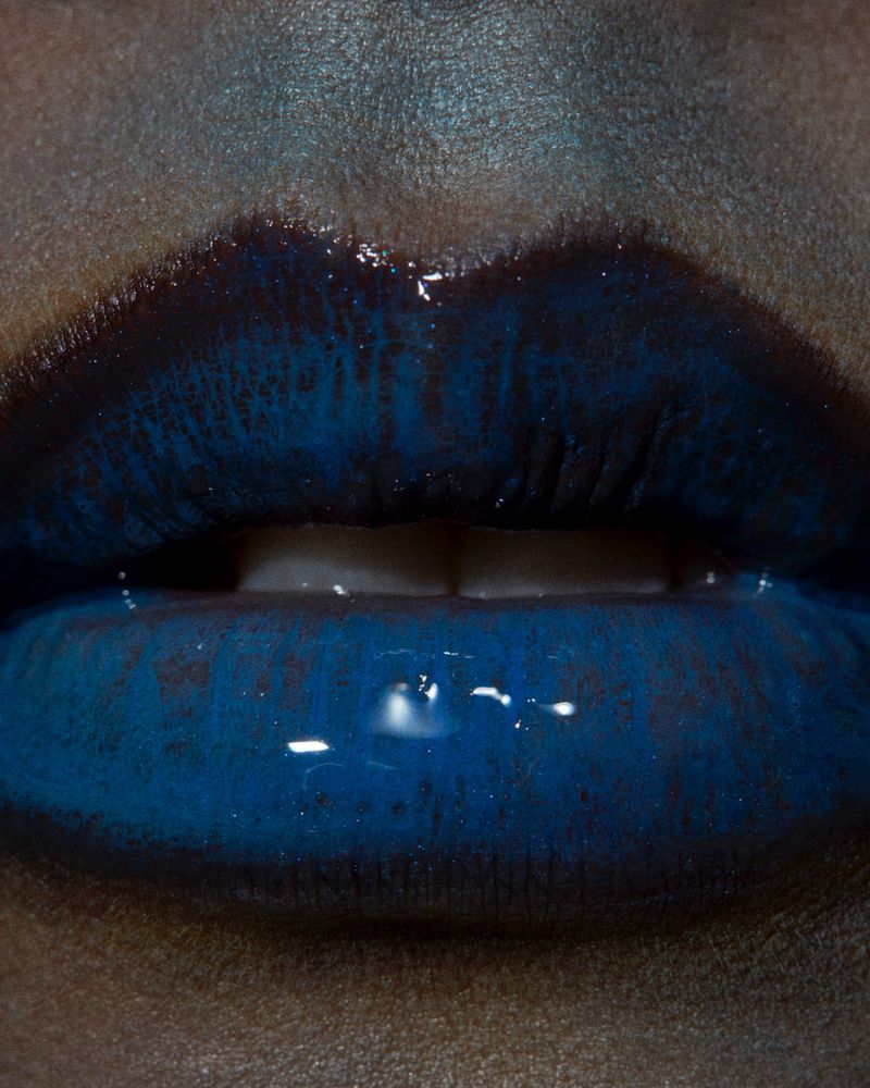 Close-up of a person's glossy, dark blue lips, set against a neutral skin tone, highlighting the rich color and texture of the makeup.