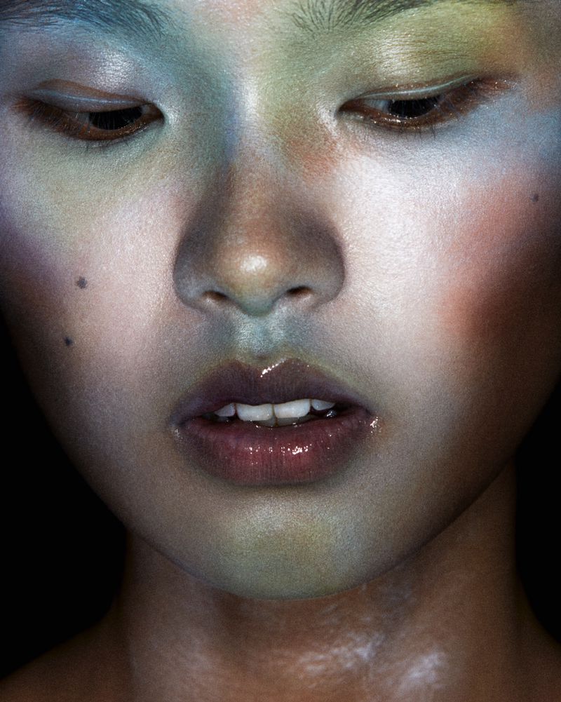 Close-up of a young adult's face with colorful makeup on face and around the eyes, set against a dark background in a beauty shot.