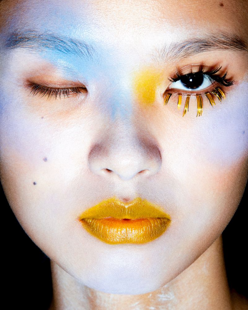 Close-up of a woman with creative makeup, including gold lipstick and embellishments around one eye, against a dark backdrop.