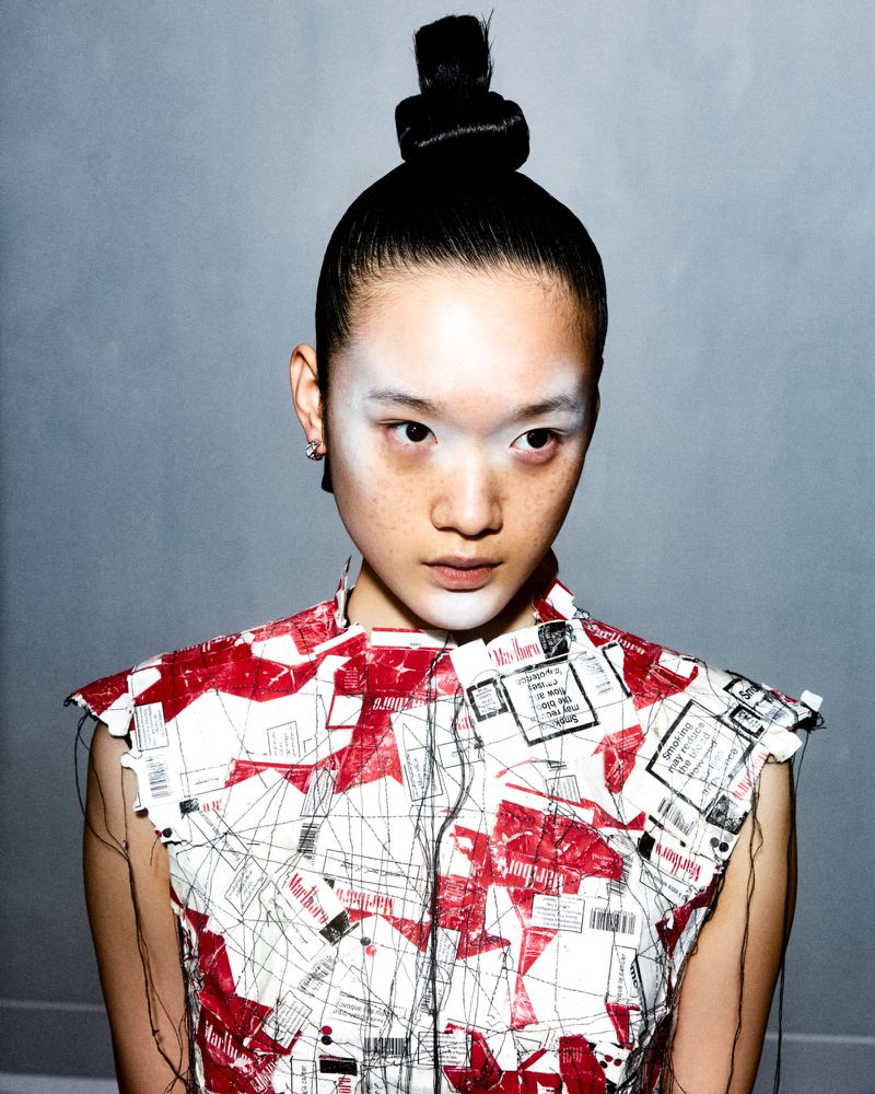 Portrait of an Asian woman with a high bun, wearing a red and white printed sleeveless top and white face makeup, against a grey backdrop.