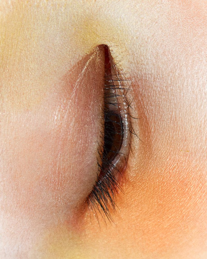 Macro photograph of a closed eye with long eyelashes, light tan skin, and soft, diffused lighting emphasizing the texture of the skin.