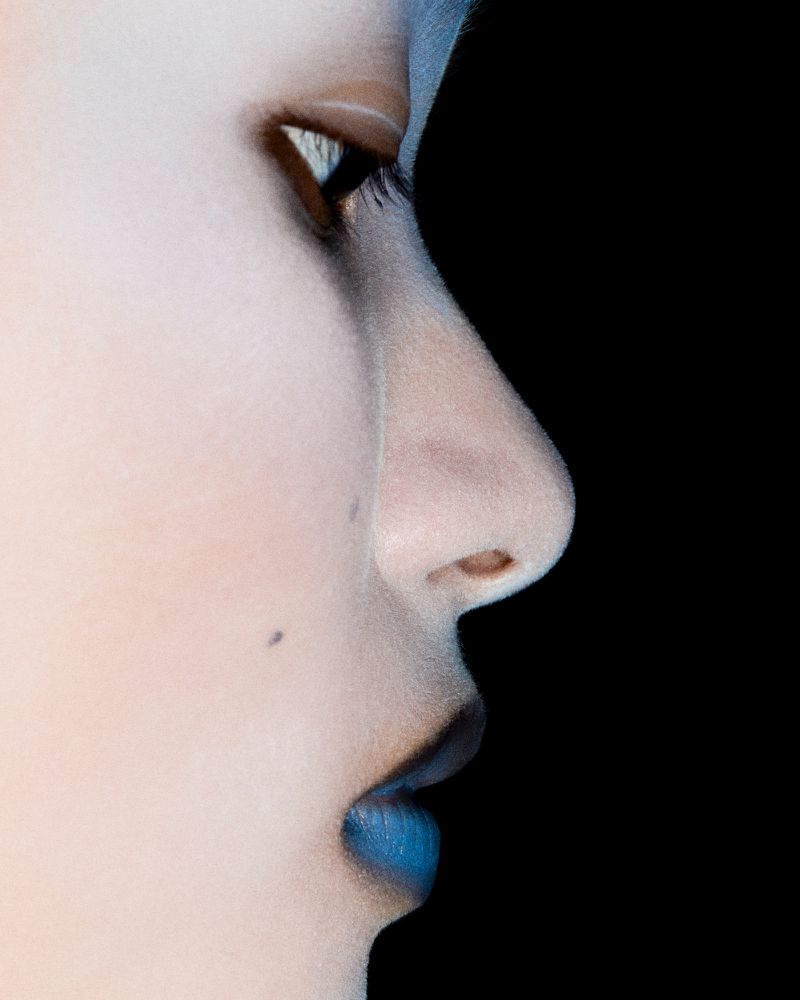 Close-up side profile of a person with light skin and blue lips against a black background, showcasing makeup and facial features.