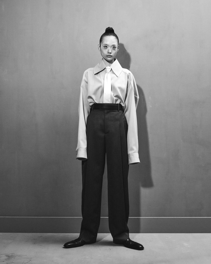 Monochrome portrait of a young adult in an oversized white shirt, tie, and dark trousers, standing against a plain wall, fashion-forward.