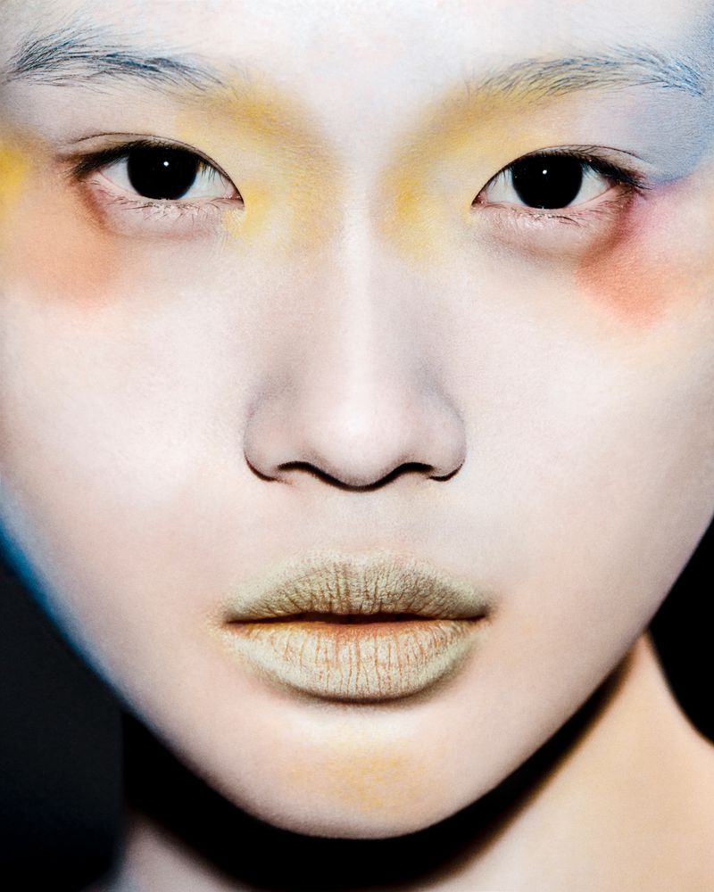 Close-up portrait of a person with artistic makeup, featuring yellow, red, and blue hues around the eyes and a unique lip color.