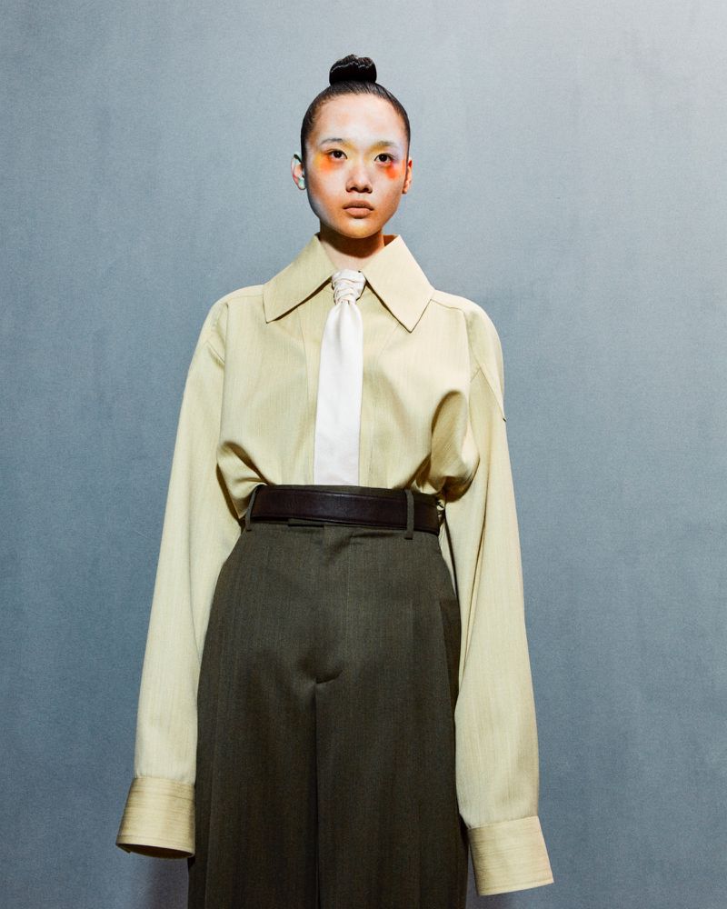 Portrait of a person with eye makeup wearing an oversized tan button-down shirt, tie, and green wide-leg trousers against a textured gray wall.
