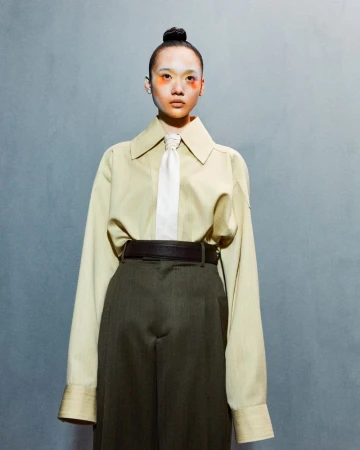 Portrait of a person with eye makeup wearing an oversized tan button-down shirt, tie, and green wide-leg trousers against a textured gray wall.