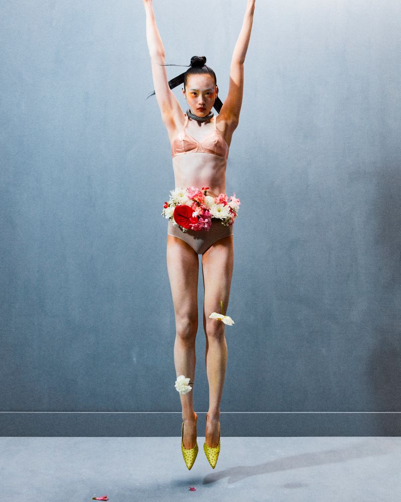Asian woman in lingerie and heels is mid-air, surrounded by flower petals against a grey wall, arms up in a studio fashion shot.