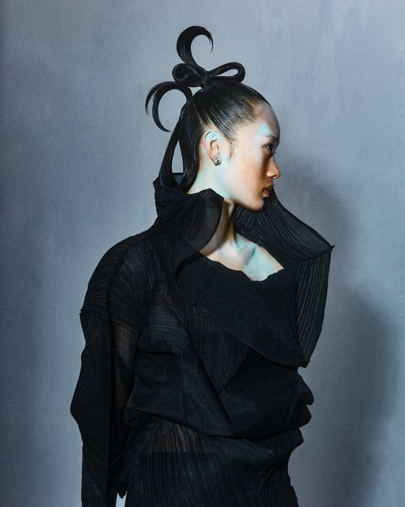 Profile of a woman with unique hairstyling, wearing a draped black outfit, in a studio setting with a cool-toned background.