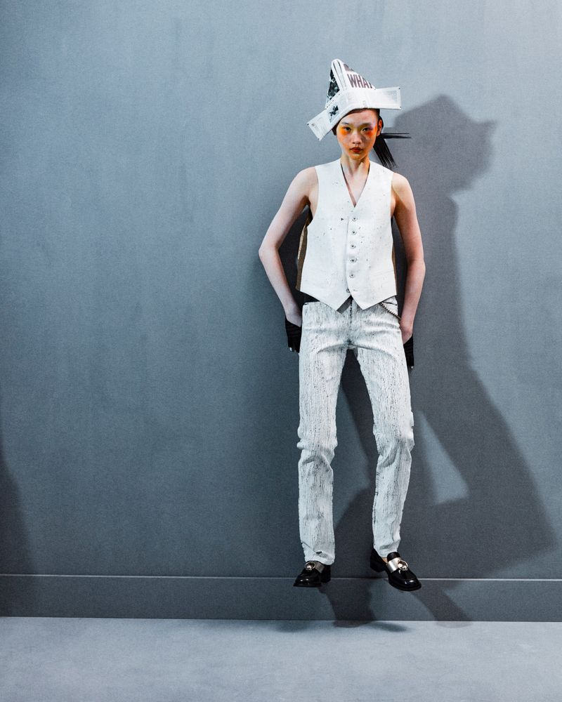 A person with orange makeup posing against a gray wall, wearing a white vest and decorative hat in a fashion-style studio portrait.