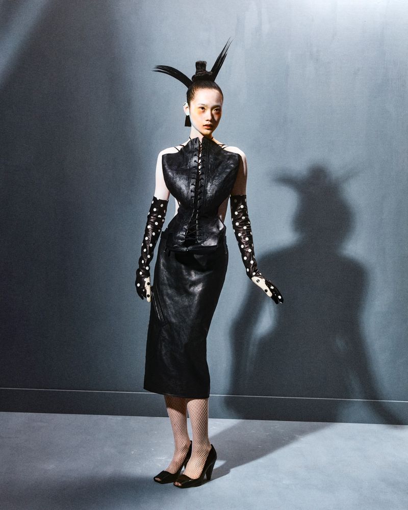 Full-length studio shot of a slender Asian female fashion model in a black dress, polka-dot gloves and high heels, set against a gray backdrop.