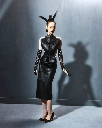 Full-length studio shot of a slender Asian female fashion model in a black dress, polka-dot gloves and high heels, set against a gray backdrop.