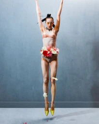 Model jumping in the air wearing a floral waist arrangement, underwear, and yellow shoes against a grey studio backdrop.
