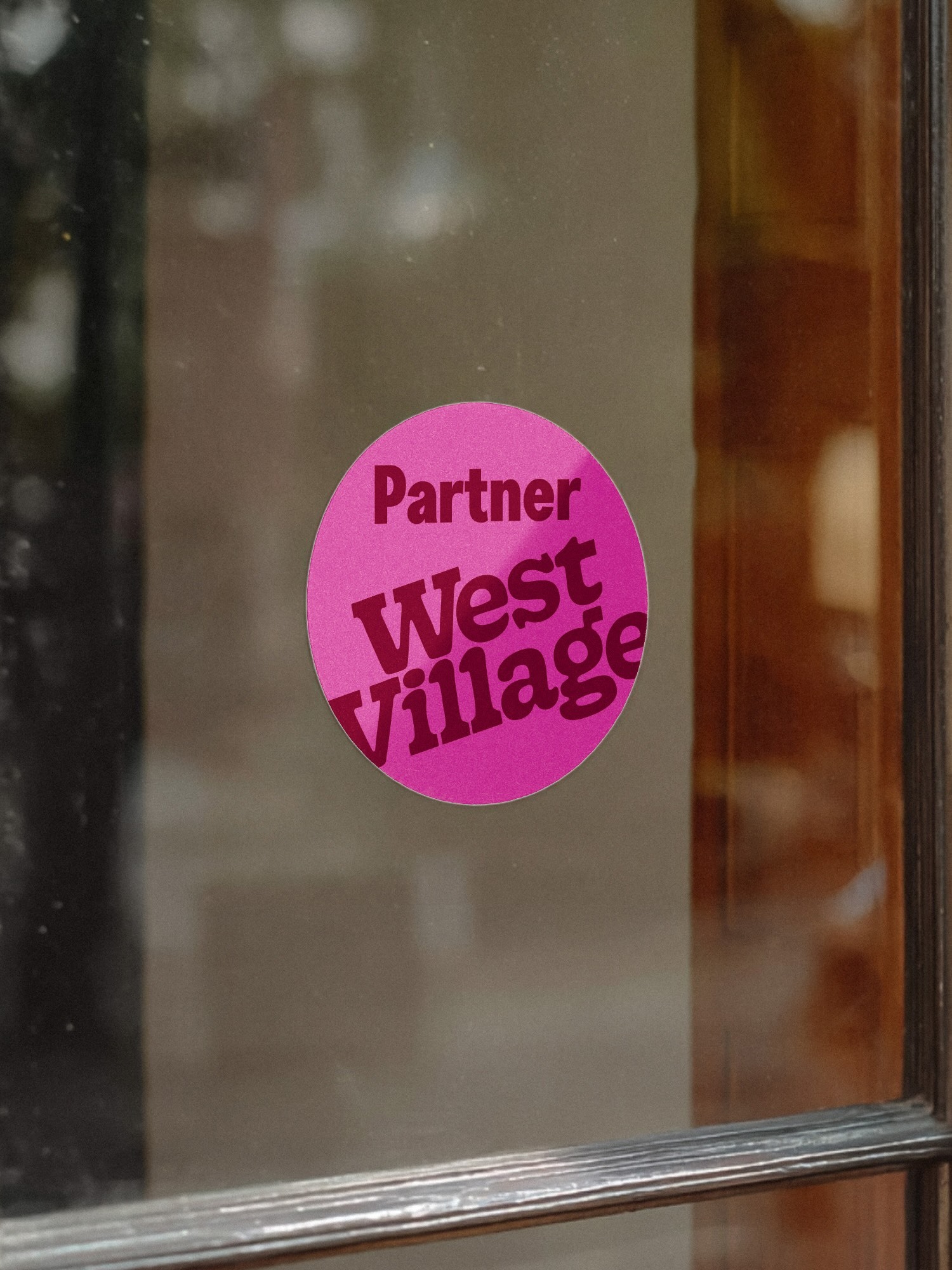 Round pink sticker with the text Partner West Village applied to a glass window pane