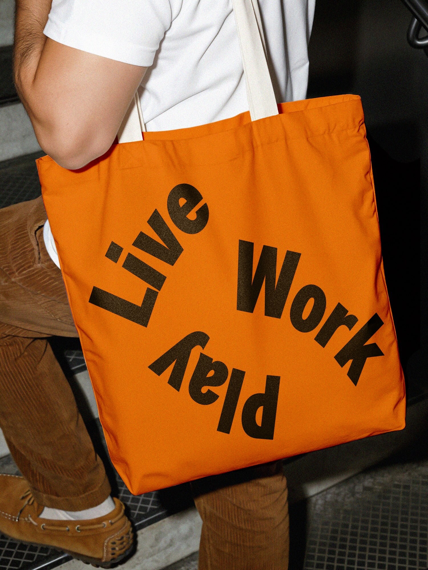 Person carrying a bright orange tote bag with bold black typographic text on the side over brown trousers.