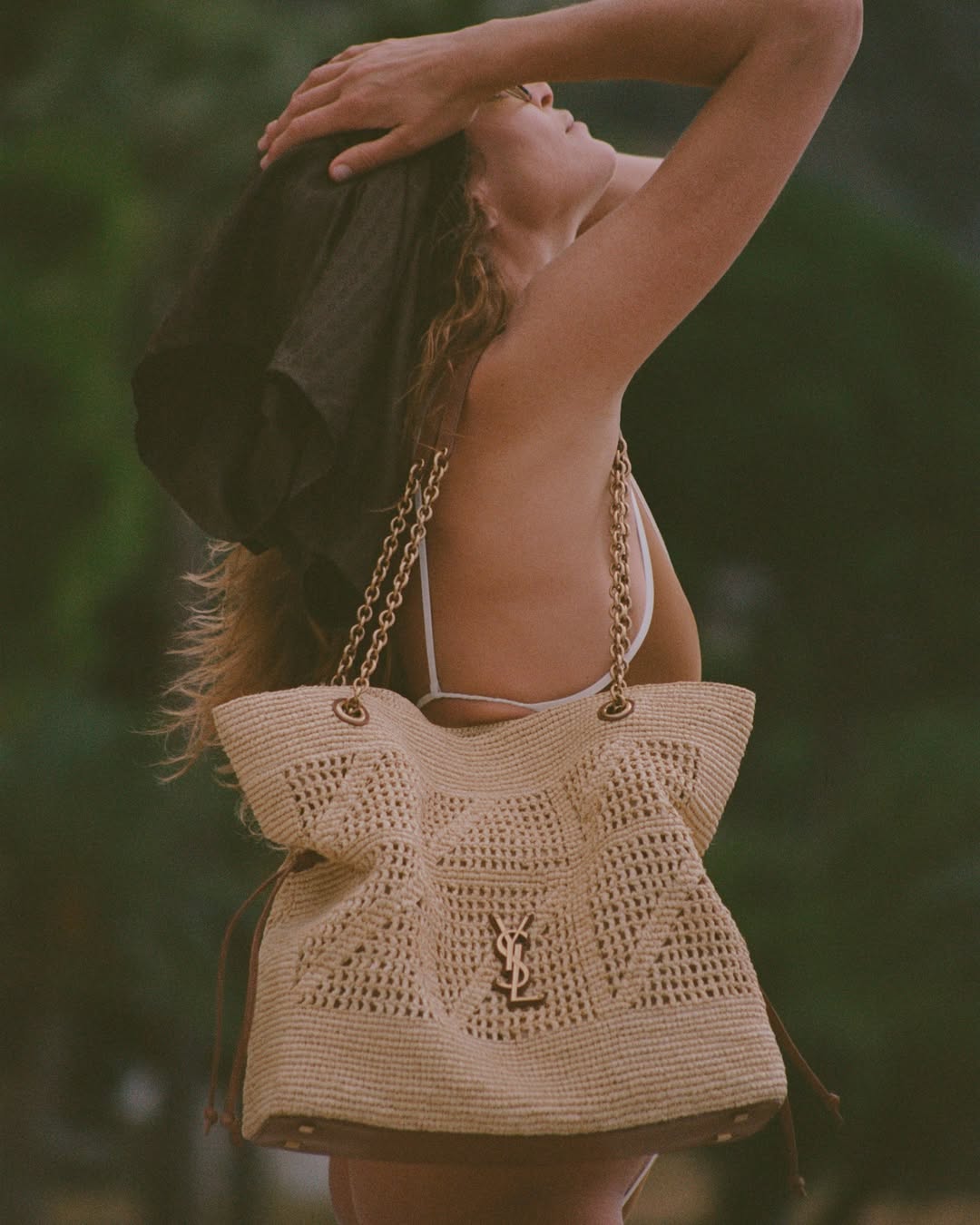 Model wearing a woven straw shoulder bag with chain straps outdoors against a natural green background