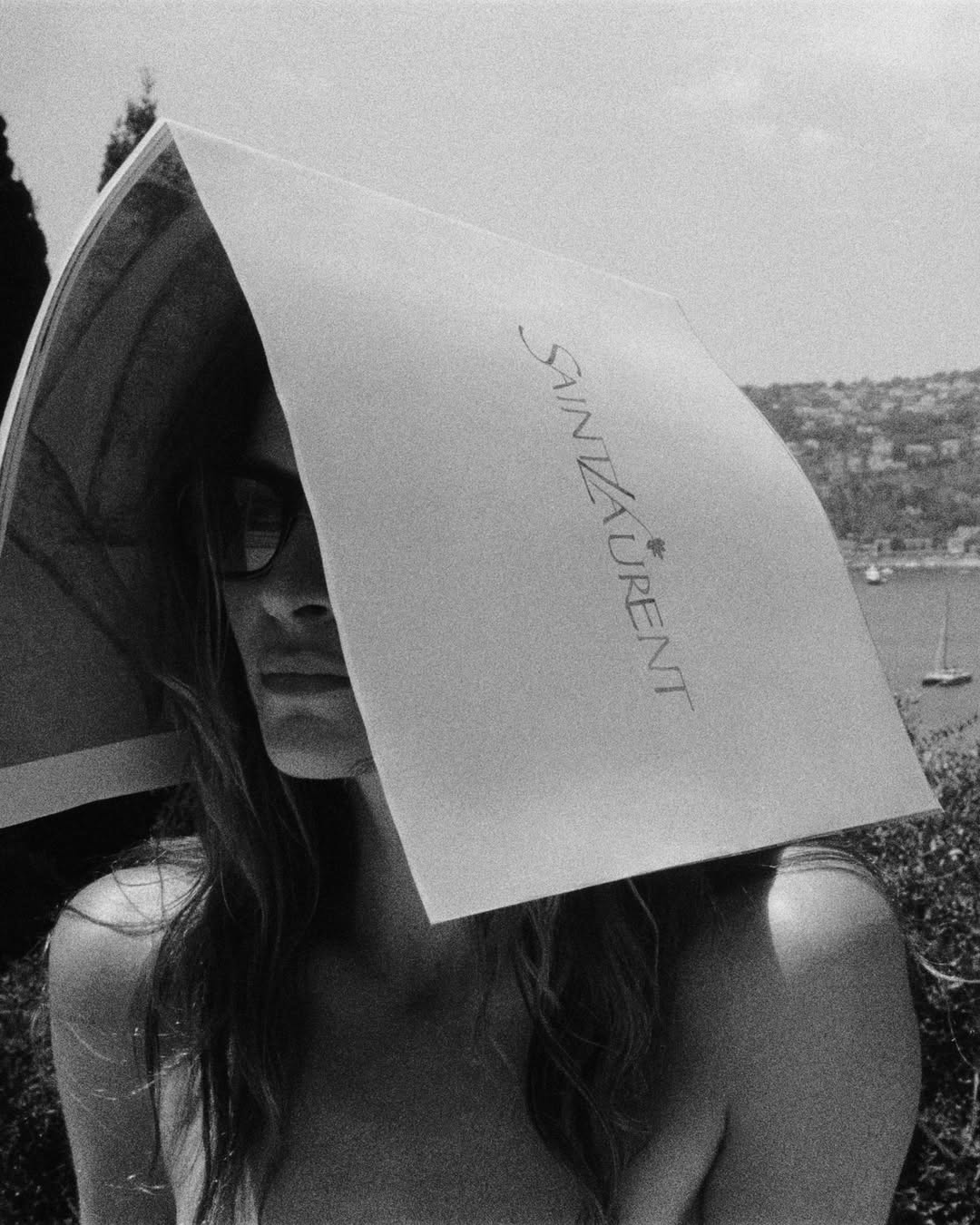 Model wearing a magazine over her head as a makeshift sun hat with visible Saint Laurent logo in a monochrome outdoor campaign shot