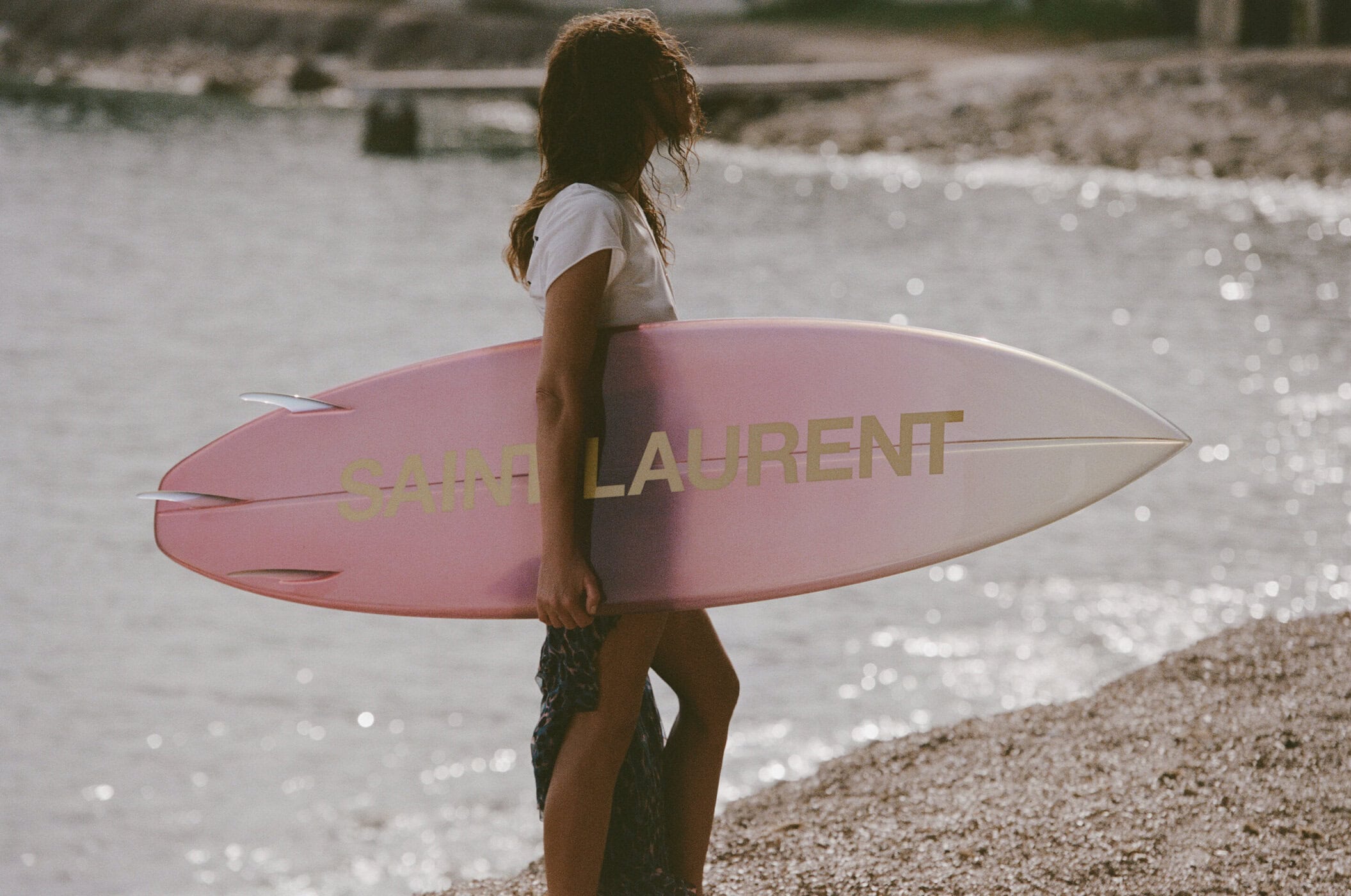 Woman holding a pink Saint Laurent surfboard with gold lettering on a pebbled beach by the water.