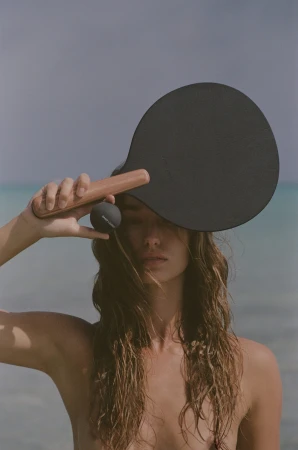 Model holding a black paddle and small ball covering her eyes outdoors against an ocean background