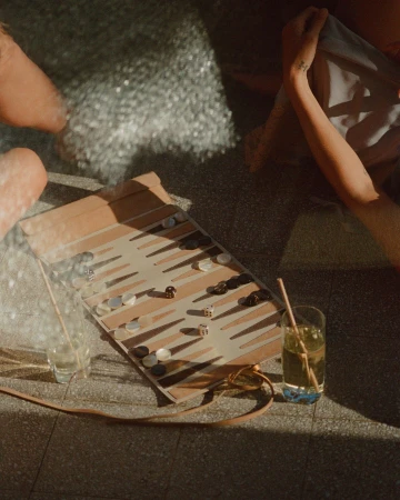 High-angle view of a backgammon board and glasses of iced drinks on a sunlit stone surface with partial figures.