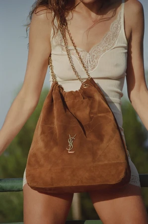 Model wearing a white lace slip dress holding a large brown suede shoulder bag with gold chain straps by Saint Laurent.
