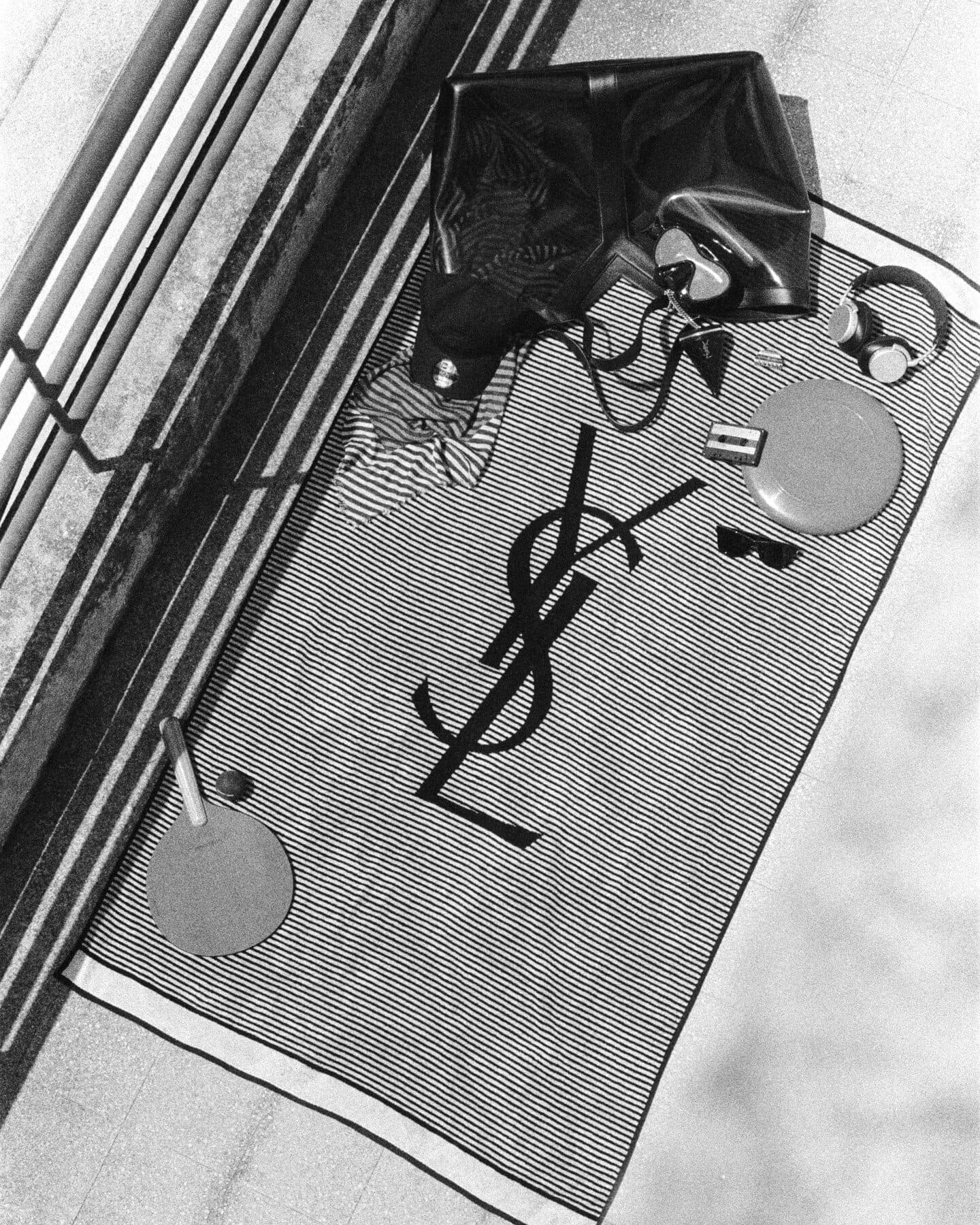 Overhead black and white photo of a Saint Laurent beach towel with YSL logo, tote bag, headphones, and ping pong paddle