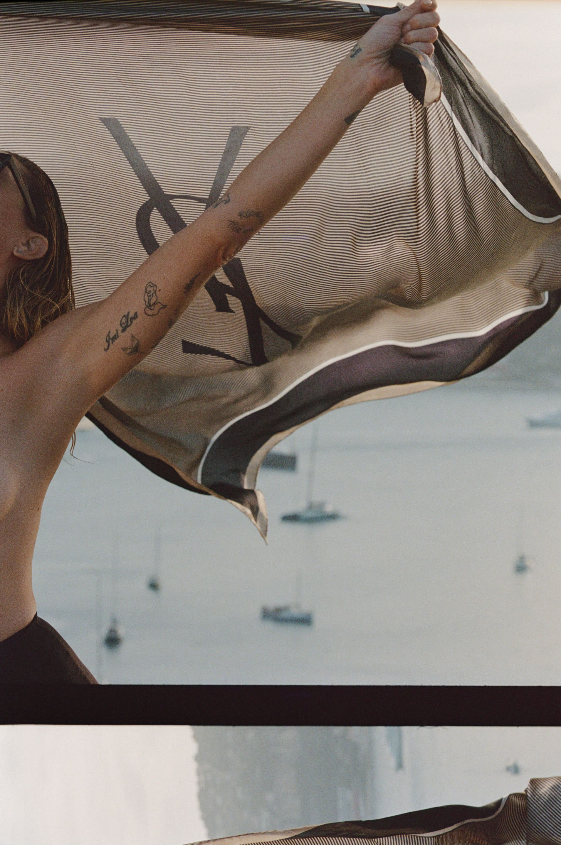 Model holding up a large Saint Laurent logo scarf with sailboats visible in the background waters of South of France.