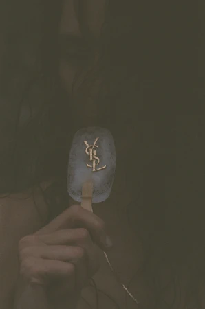 Close-up of a person holding an ice pop embedded with a gold YSL logo for Saint Laurent.
