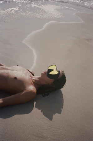 Model lying on a sandy beach wearing a diving mask during the Saint Laurent Shades of Summer campaign.