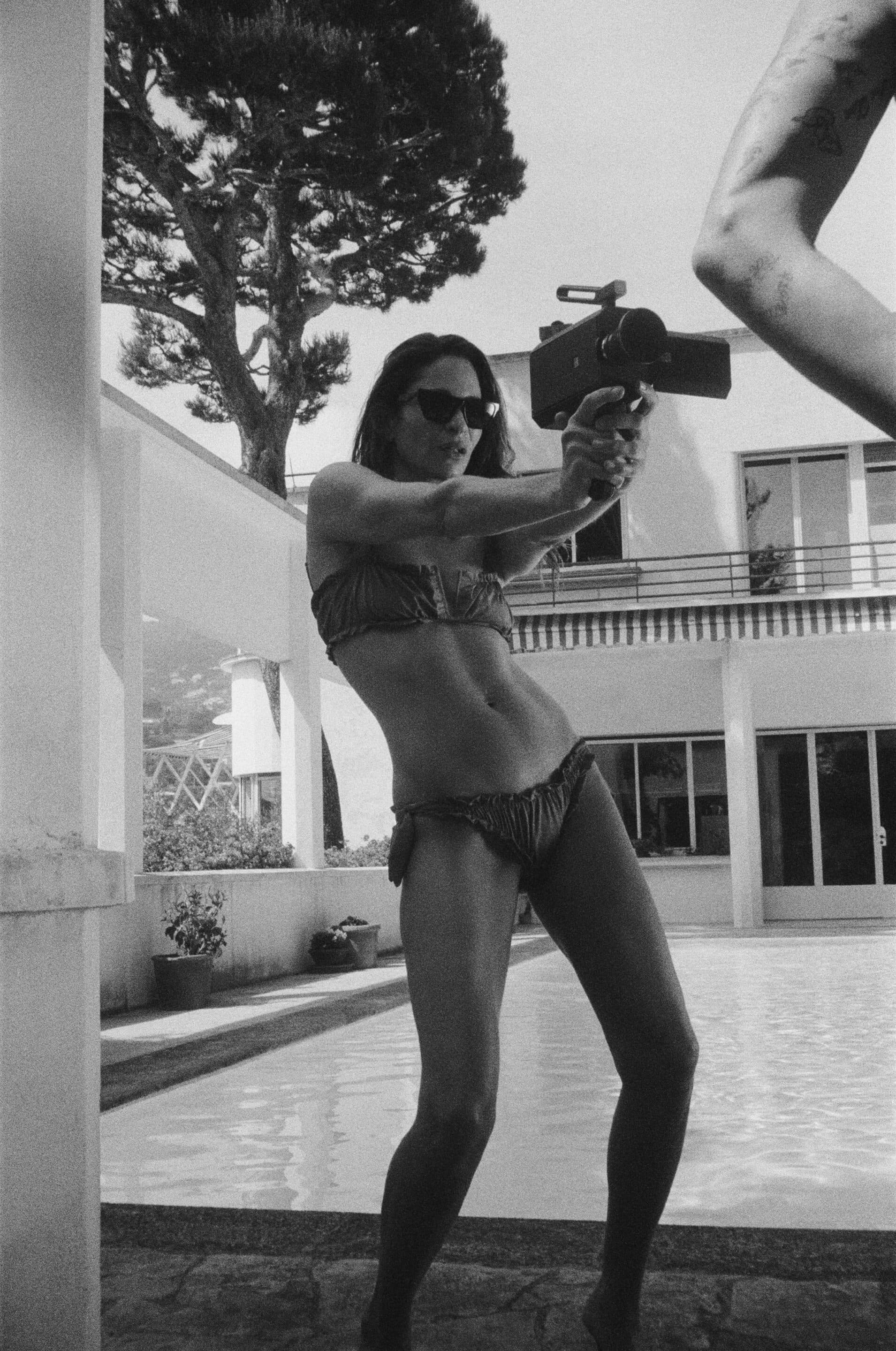 Monochrome image of a woman in a bikini holding a vintage movie camera beside a pool for Saint Laurent.