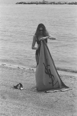 Model standing on a sandy beach holding a branded Saint Laurent towel with calm water in the background.