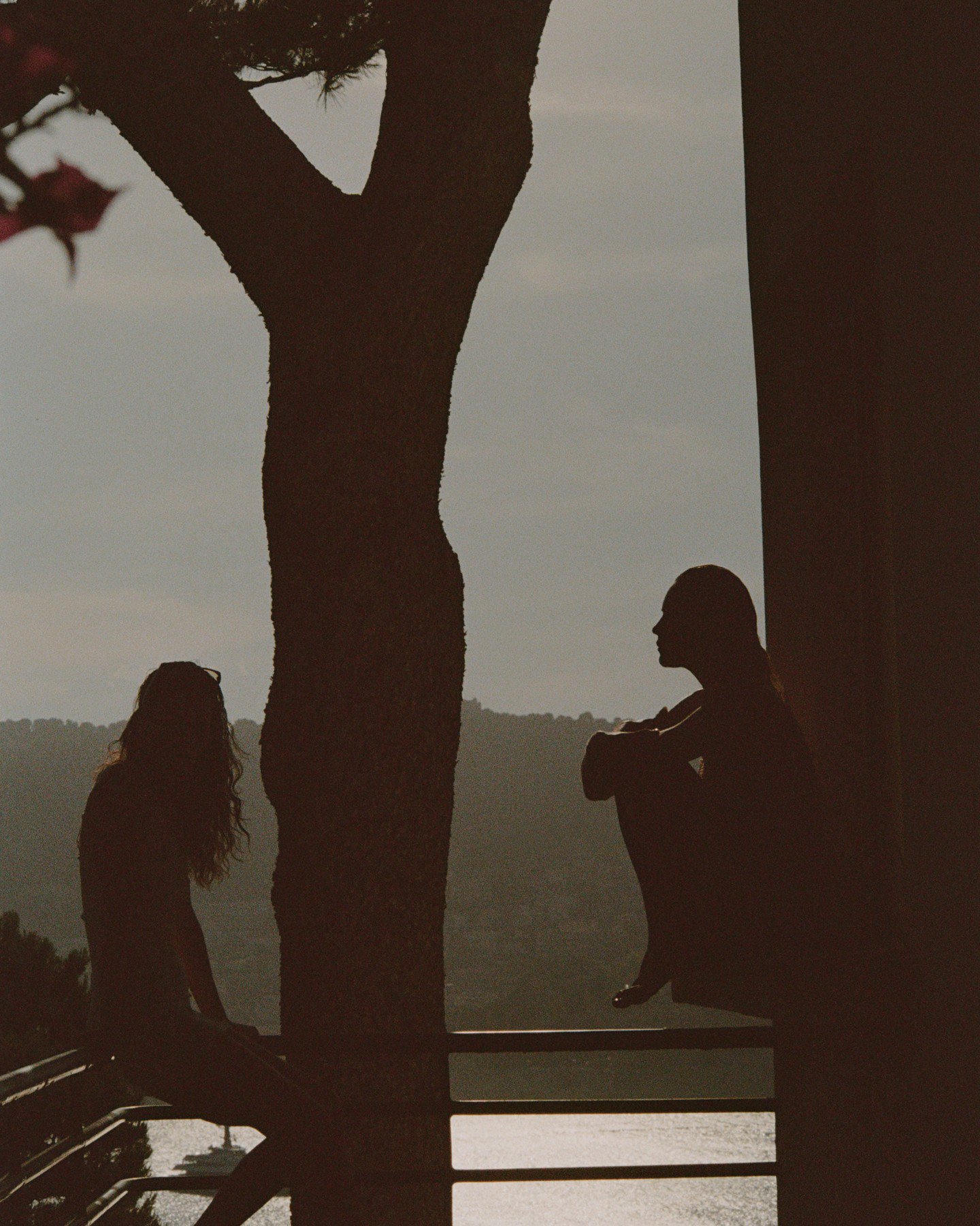 Silhouettes of two models seated on a balcony railing outdoors against a pale sky and distant hills