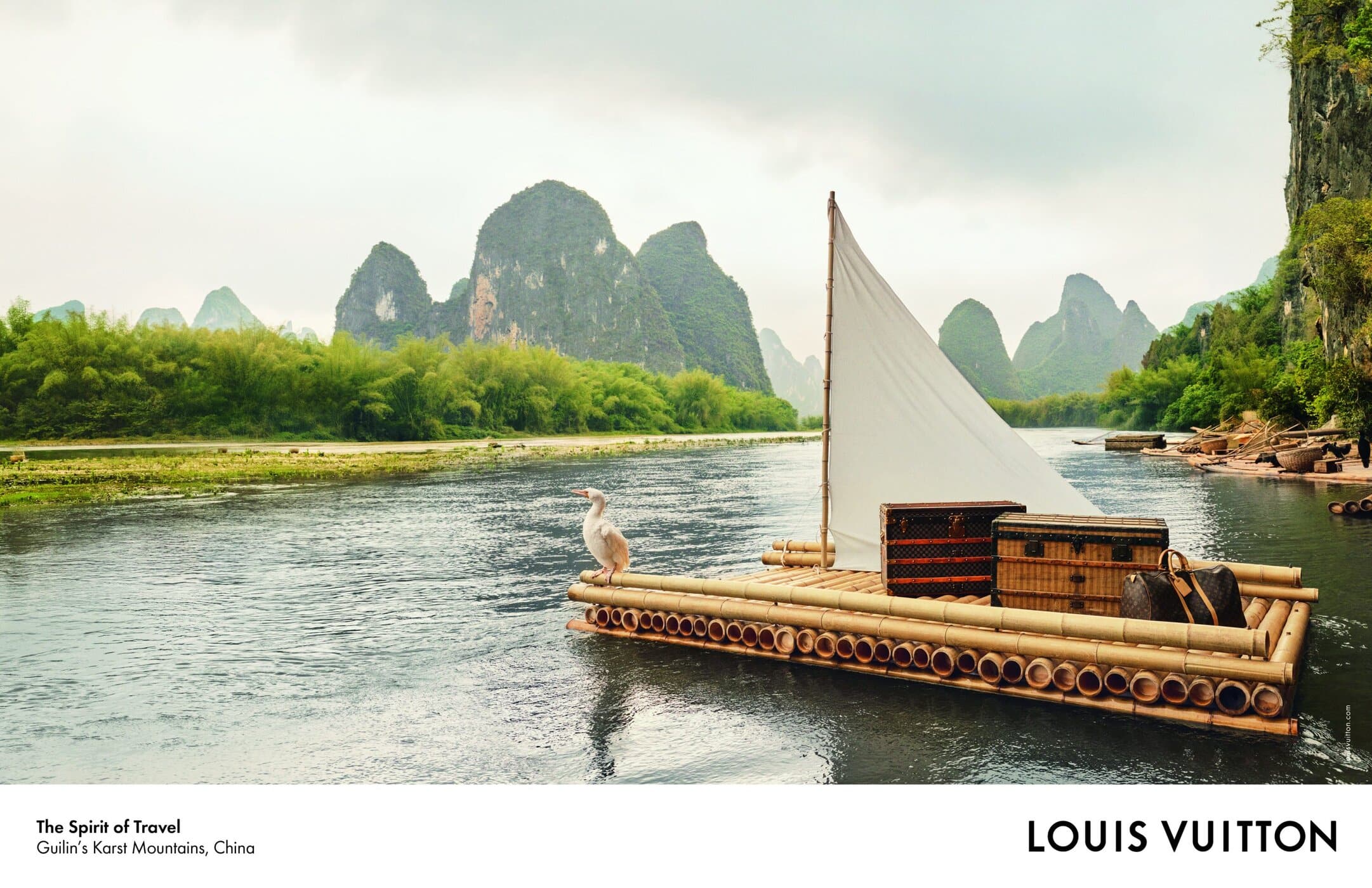 A wooden raft carrying Louis Vuitton luggage and a white-sailed mast floats on a river surrounded by limestone karst mountains.