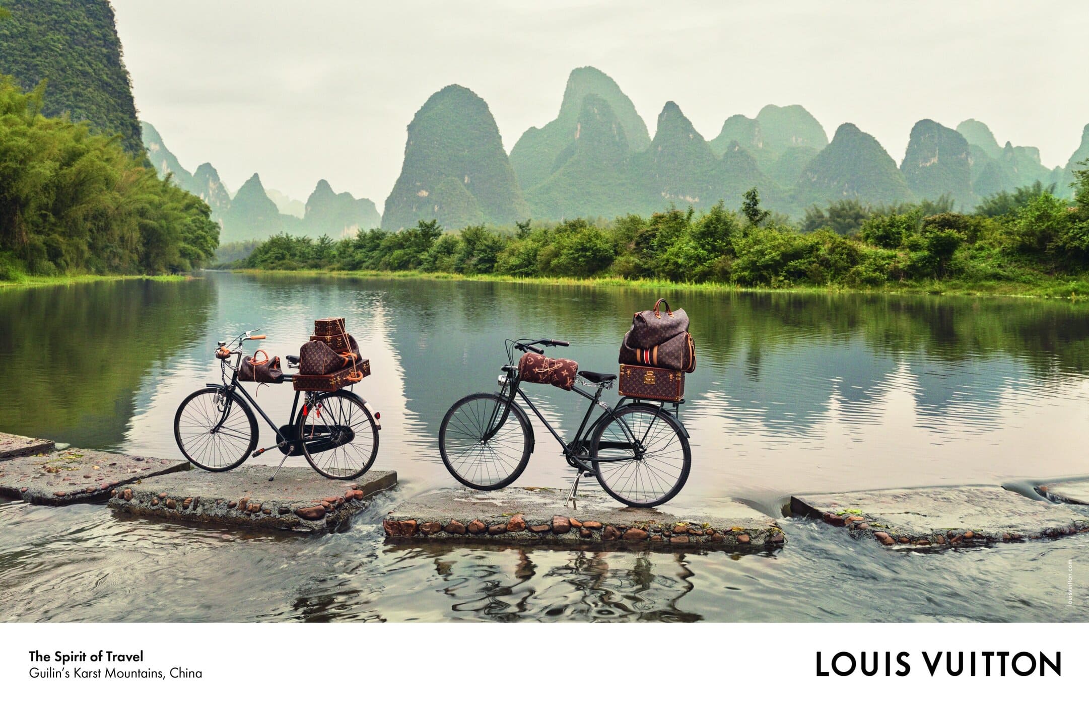 Two bicycles loaded with Louis Vuitton luggage parked on stone paths across a river with karst mountains in China.