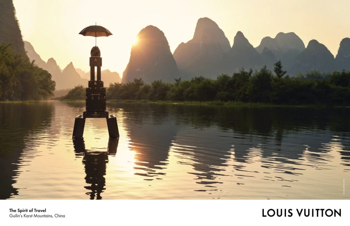 Stacked monogram luggage standing in a calm river beneath towering limestone karst mountains at sunset.